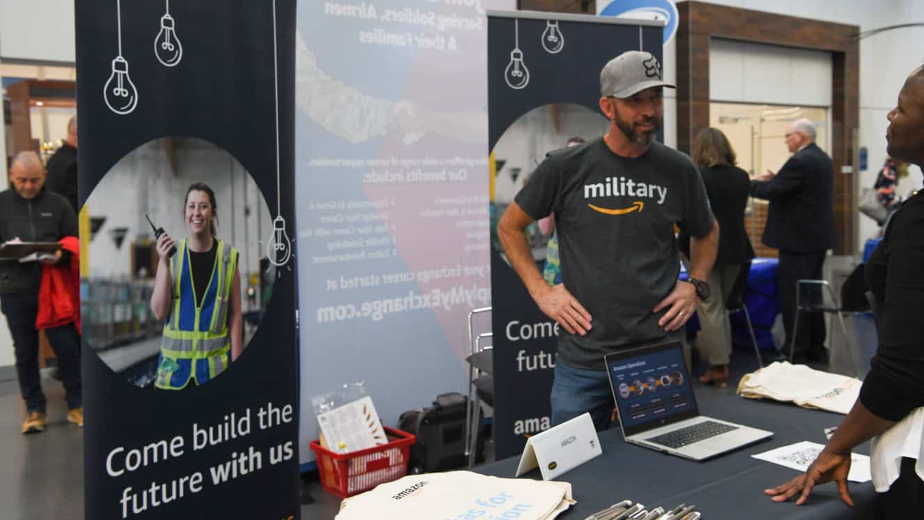A representative from Amazon talks to job seekers at the Army Community Service Job Fair, where companies are hiring military members for roles and offering remote jobs for veterans.