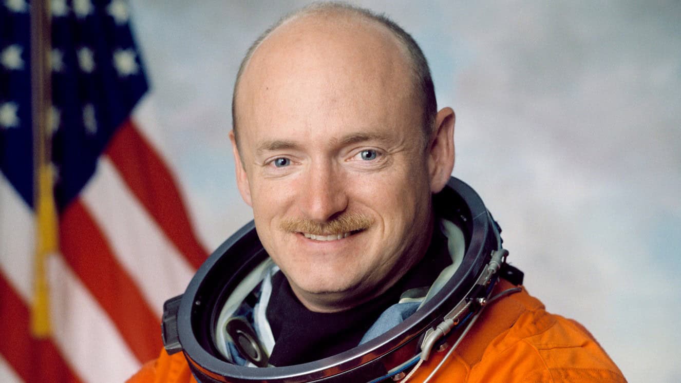 Official Portrait of Astronaut Mark E. Kelly. Kelly is wearing the orange Launch and Entry Suit (LES) with an American flag in the background.